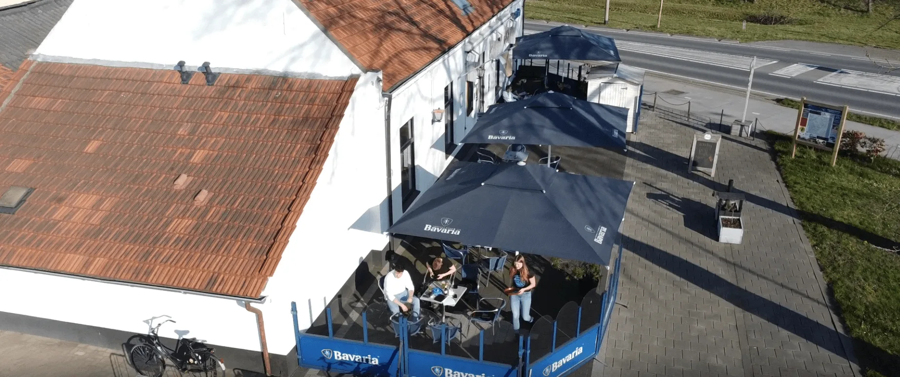 Luchtfoto van Café De Statie in Maasbracht: het terras met blauwe parasols naast het witte pand met rode dakpannen