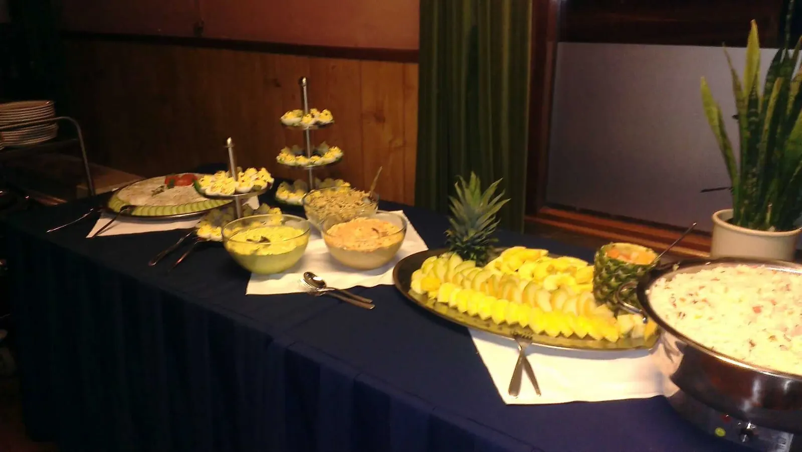 Koud buffet op een blauw gedekte tafel: etagères met gevulde eieren, schalen salade en een schaal fruit met ananas
